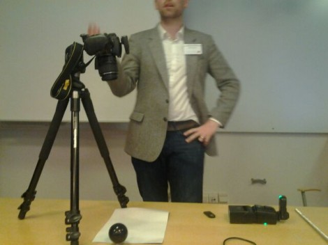 Gareth stands behind a table in a lecture room. In front of him is a camera on a tripod. The camera is set up to take a R.T.I. and so there is also a flash gun, a pool ball and a remote control on the table. 
